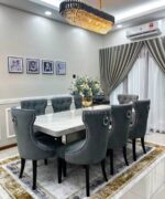Transform your dining space with the luxurious Godwin Series IX Premium 8 Seater Dining Set. This elegant dining table features a stunning marble top, paired with plush gray tufted chairs for ultimate comfort and style. Perfect for modern homes, this sophisticated set is beautifully complemented by a statement chandelier, stylish curtains, cherished family photos, and a vibrant floral centerpiece—creating a warm and inviting atmosphere ideal for gatherings and celebrations. Upgrade your home décor today with the Godwin Series IX marble dining set for 8 and experience the perfect blend of luxury and functionality in your modern dining room.