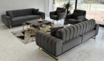 Elevate your modern living room with the luxurious Glory Series IX Premium Fabric 7-Seater Sofa Set in elegant gray. This contemporary space features stylish gold-accented coffee tables, a lush indoor plant positioned beside expansive windows draped with white curtains, and stunning marble flooring that adds a touch of sophistication. Perfect for those seeking upscale comfort and timeless design in their home decor.